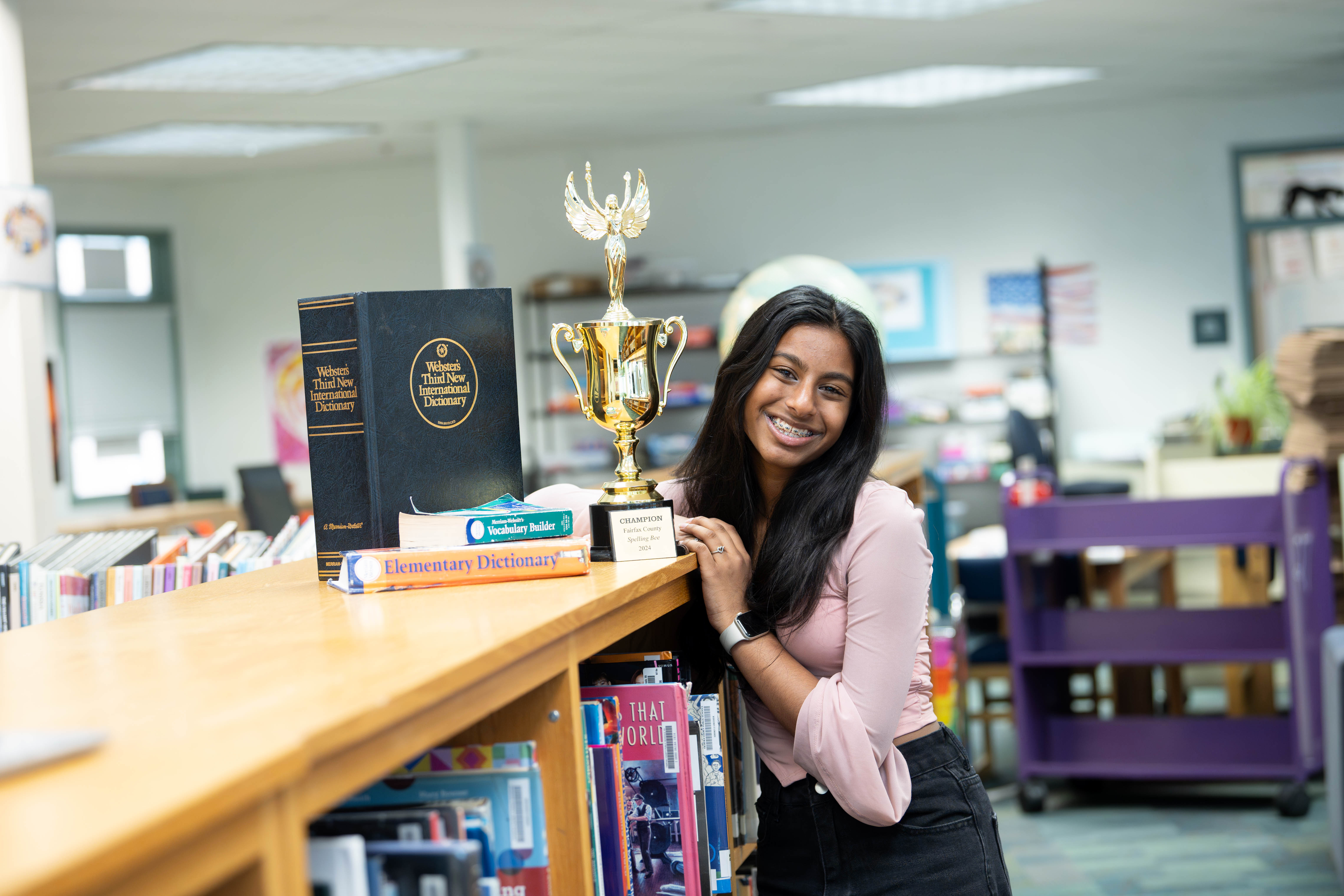 Rachel Carson seventh grader Ankita Balaji smiles as she leans on a short bookshelf. On top of the bookshelf are three dictionaries and a trophy.