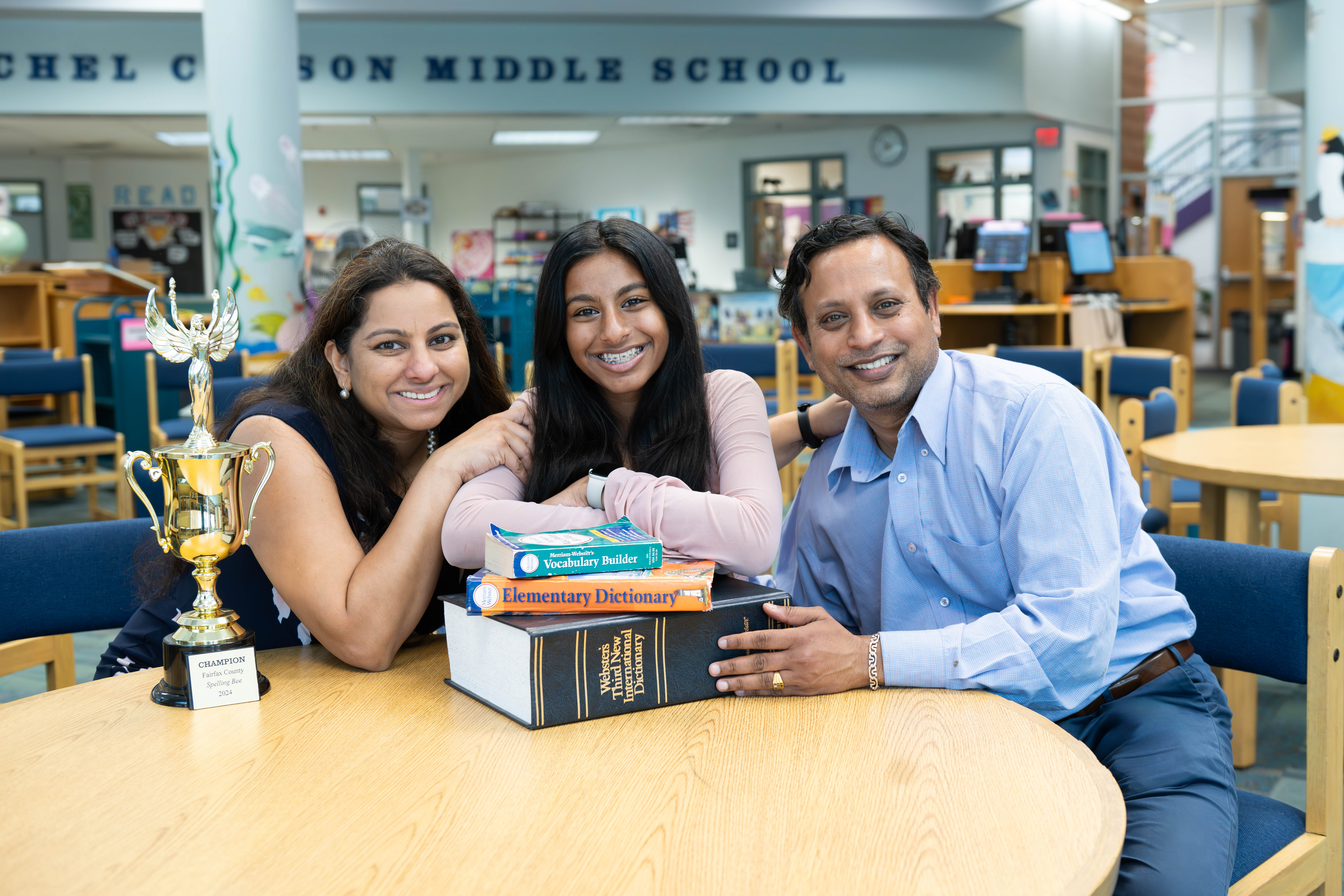 Ankita Balaji poses with her parents, mother on the left, father on the right. The three of them are seated at a table in the Rachel Carson Middle School library. Stacks of dictionaries and Ankita's trophy are on the table in front of them.