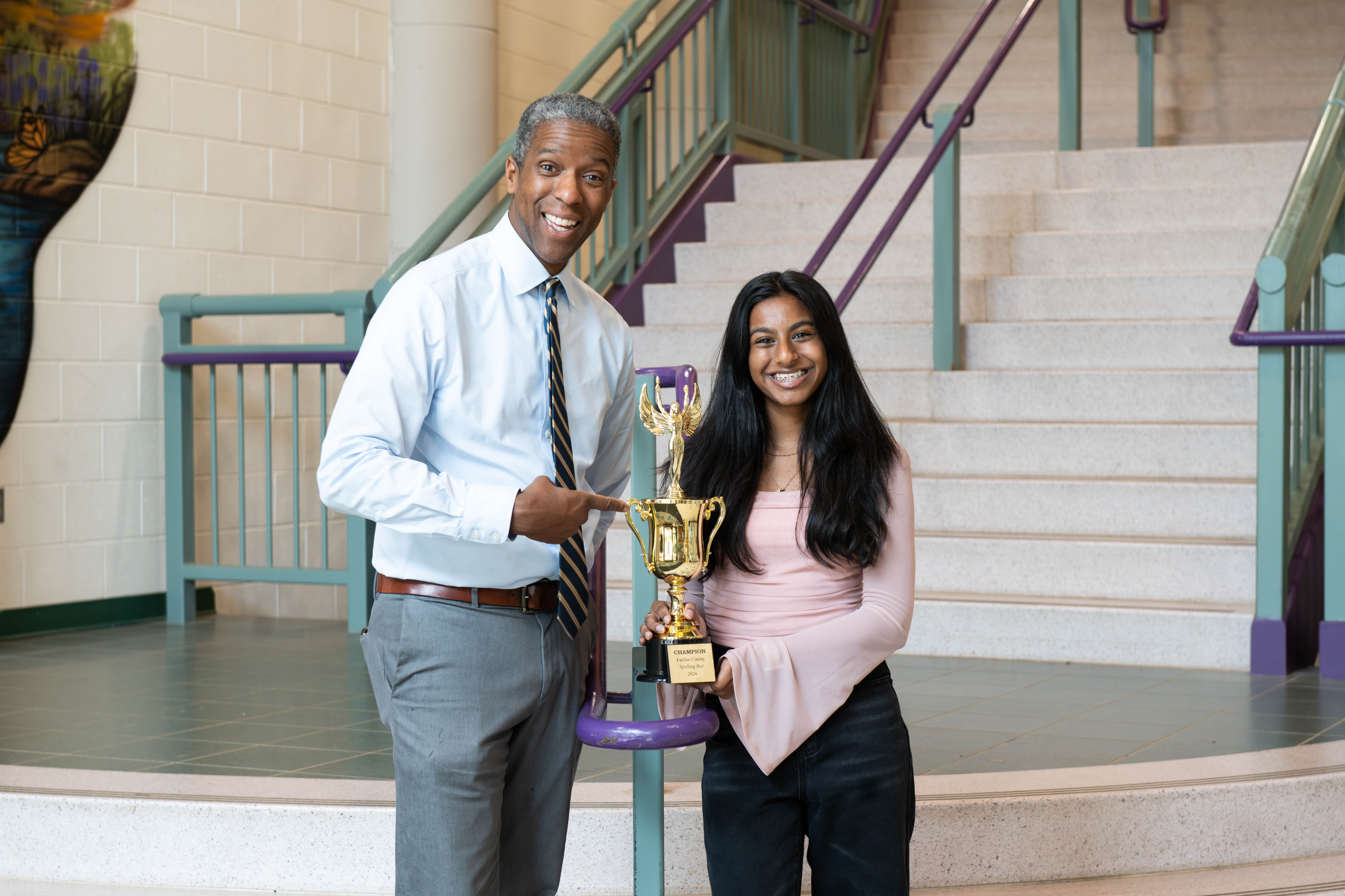 Ankita stands in the main foyer at Rachel Carson Middle School. Stairs with purple and green railings are behind her. Ankita holds her trophy. Standing next to Ankita, pointing at the trophy and smiling, is Carson Principal Gordon Stokes.
