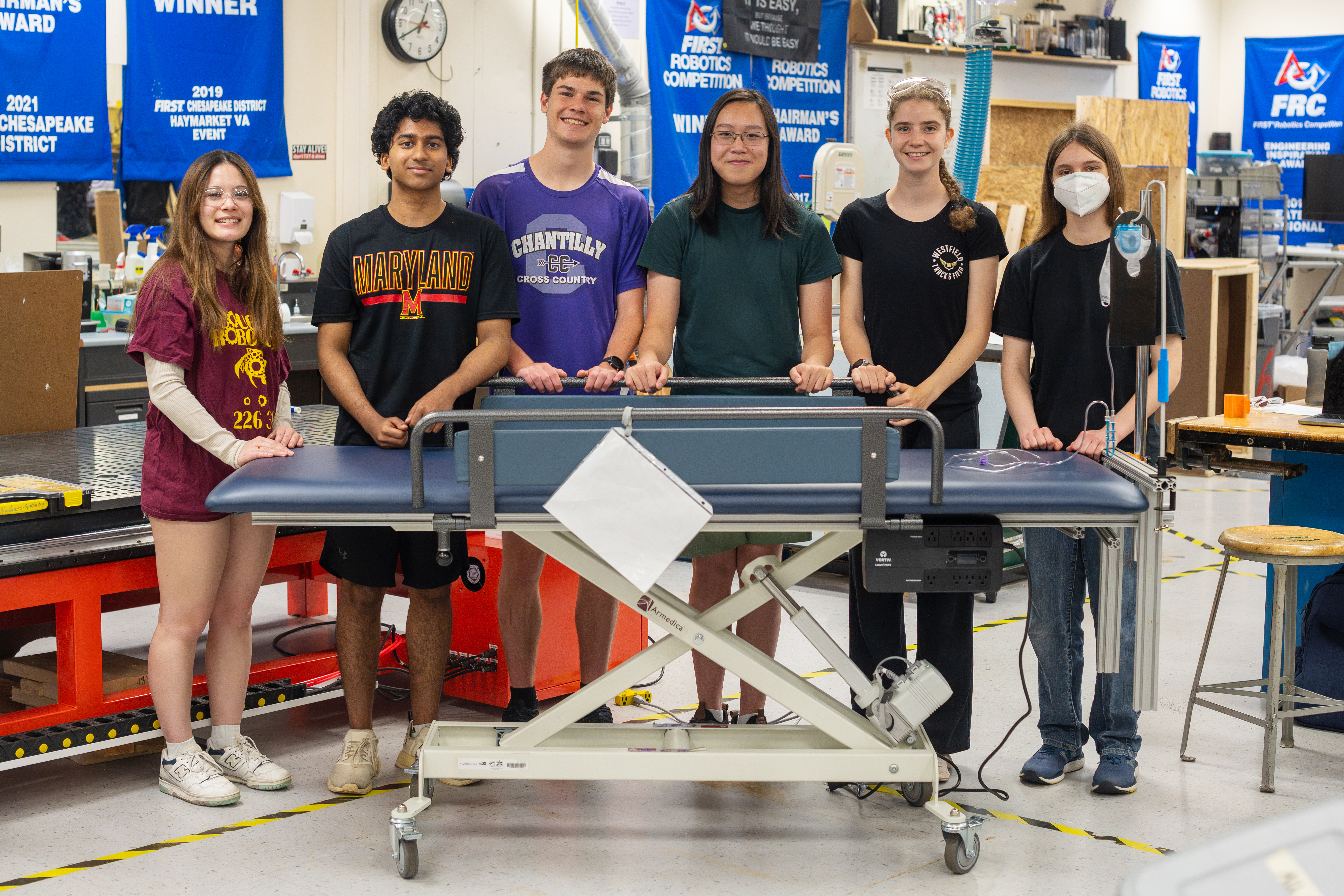 The six students on the Engineering team smile as they stand behind the bed.