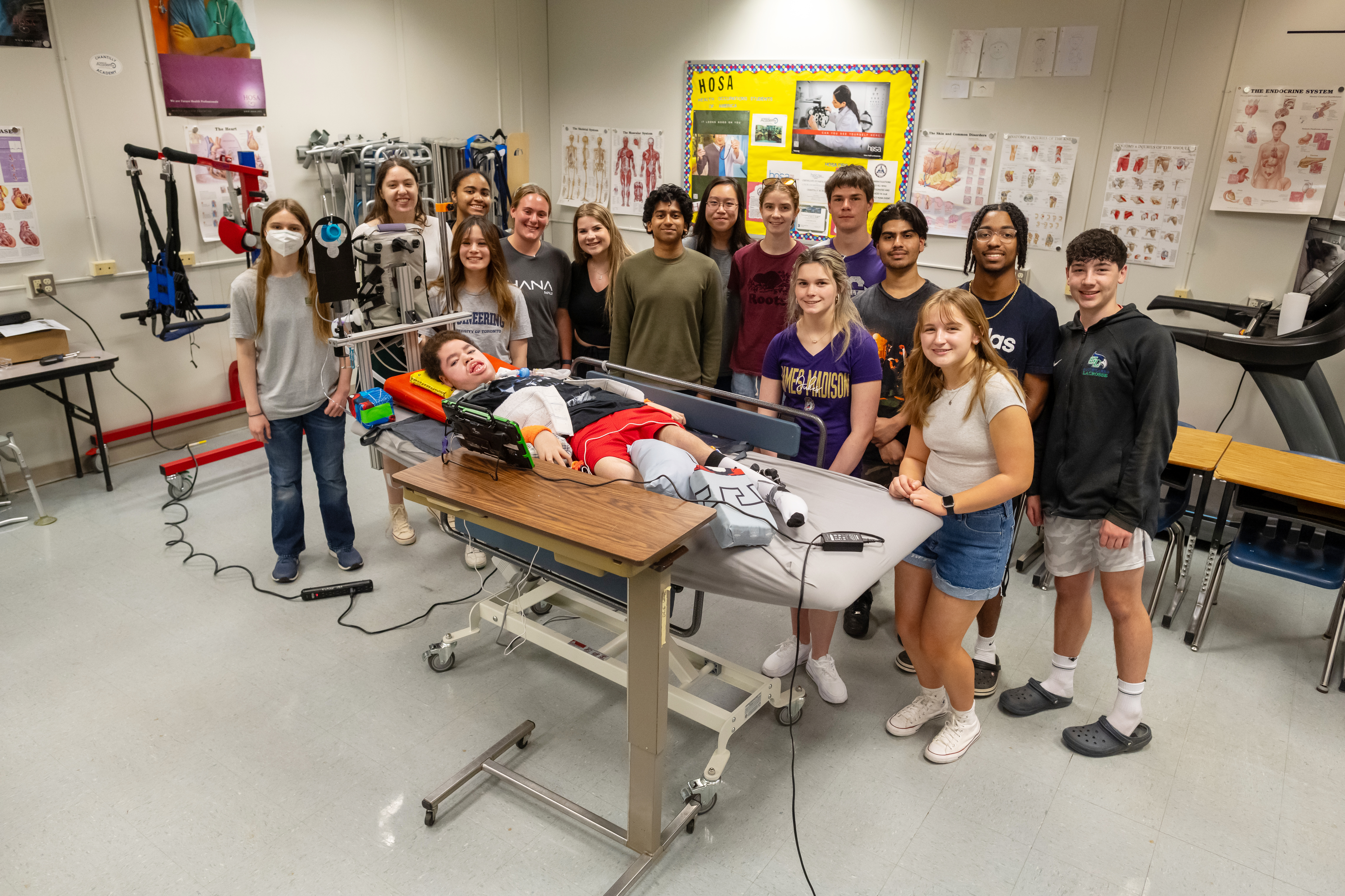 The Chantilly Academy Engineering and PT/OT students  stand and smile behind Liam in the PT/OT classroom. Liam, in the bed, is looking toward the camera. A power strip runs from one wall to the bed.