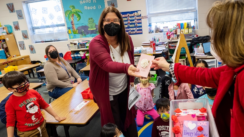 2 adults in a classroom with children, one presenting a Valentine's day card