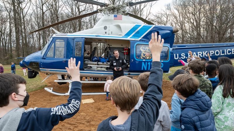 Students raise their hands while a man in uniform answers questions standing by a helicopter