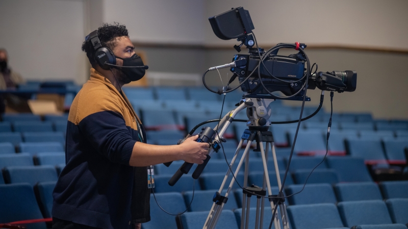Man standing behind video camera in an auditorium.
