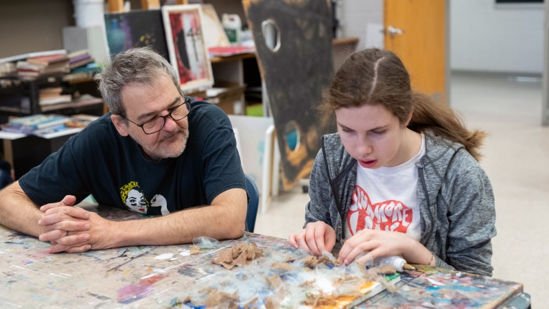 Teacher Matt Ravenstahl watches student Nora Patterson as she glues fabric to canvas.