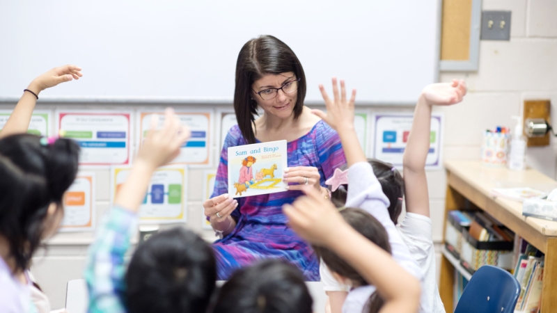 A teacher reads to students who have their hands raised. 