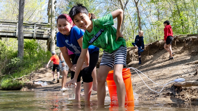 A student smiles as he lowers a cup containing a young trout into the stream he's standing in.