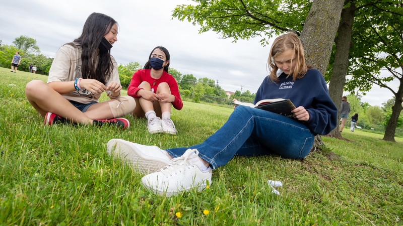 Three female students sit outdoors in the grass talking and reading. 