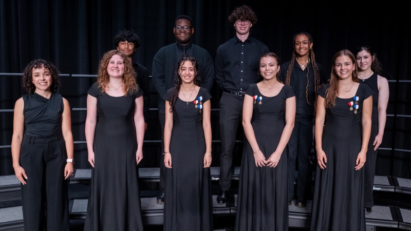 A music teacher and 8 students stand on risers before a performance. 