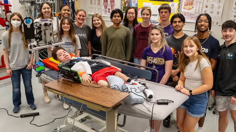 Students in Chantilly Academy's Engineering and PT/OT classes pose, smiling, with Liam Bruen, who is laying on the medical bed that the students worked to design and build.