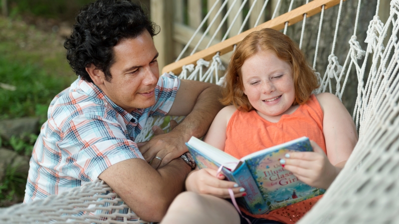 A girl reading in a hammock with her dad 