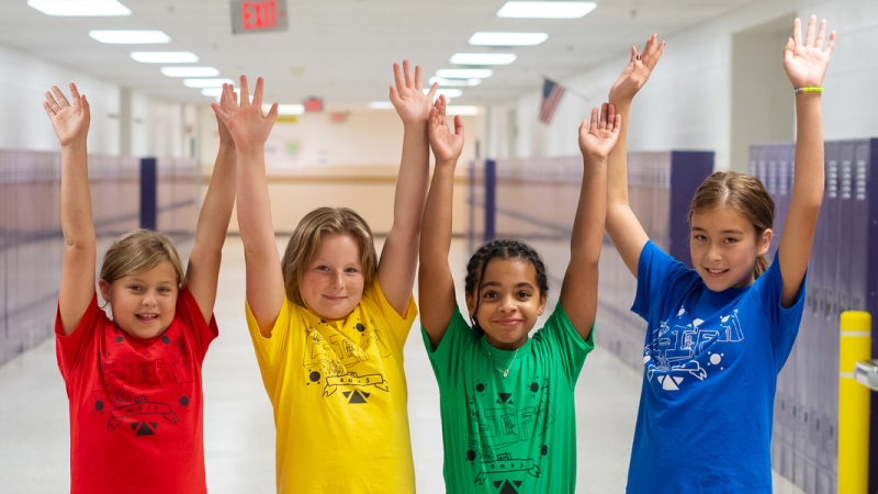 Four students in a rainbow array of colored shirts raise their hands in cheers in a school hallway.
