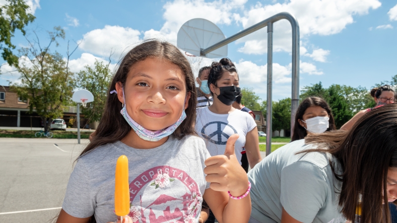 Elementary school age girl holding an orange popsicle and flashing a thumbs up. 