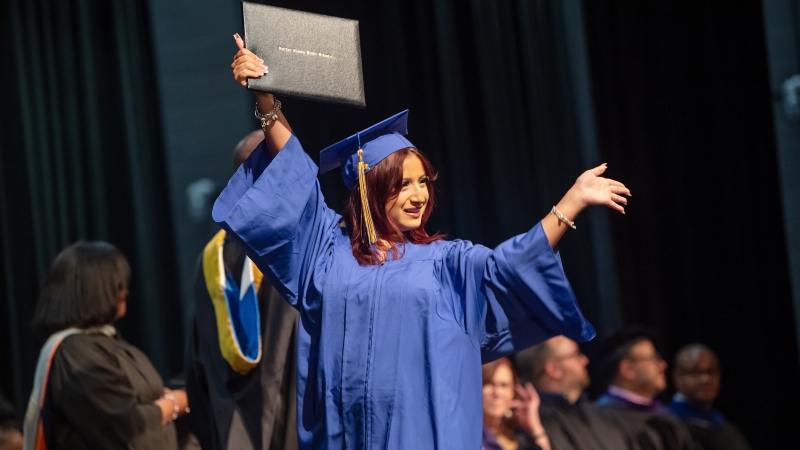 A girl in a graduation robe holds up her diploma