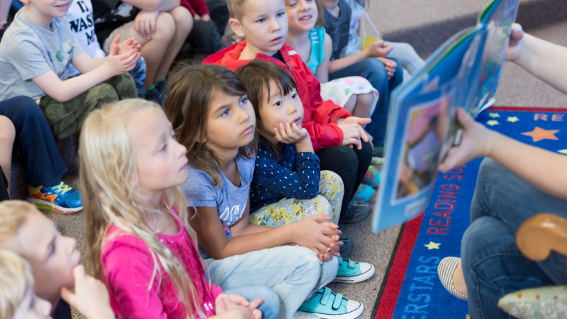 a teacher reads aloud a book to young students sitting on a carpet