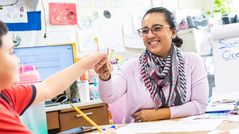 Teacher working with student at teacher desk bumping fists in celebration