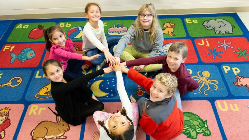 Students sitting on a classroom rug with their right hands stacked on top of each other.