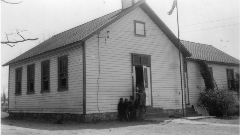 Black and white photograph of the Falls Church Colored School taken in 1942. Several students are pictured standing next to the entrance of the building.