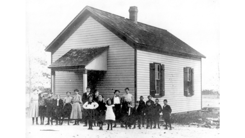 Black and white photograph of the Fairview one-room schoolhouse. It is a class photo with a large group of students and their teacher standing in front of the building.