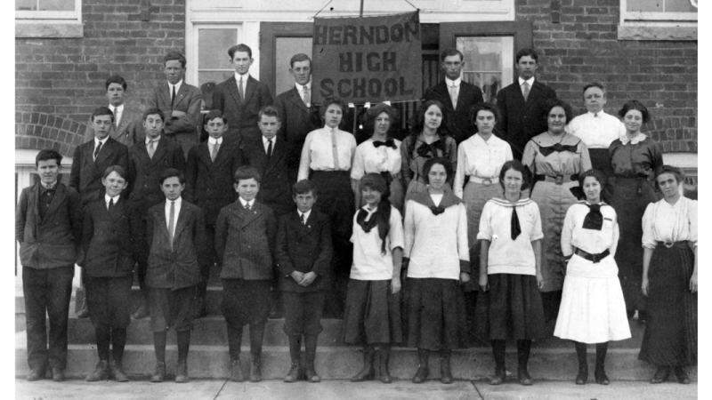 Photograph of students and teachers at the first Herndon High School. They are standing in three rows on the steps in front of the building.
