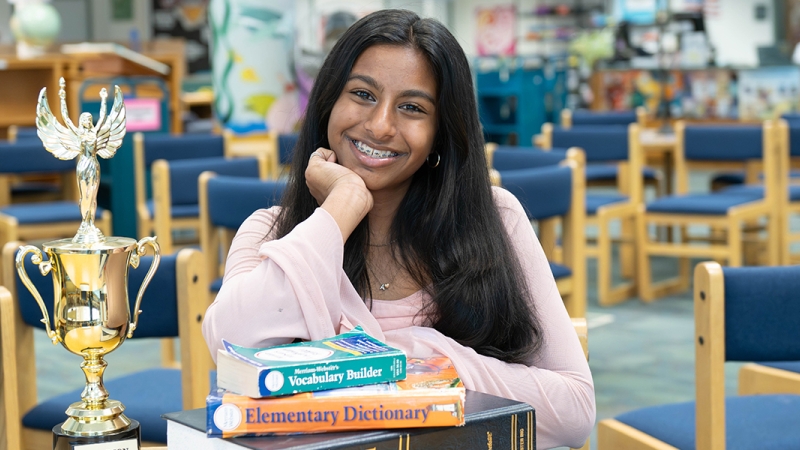 Carson Middle School seventh grader Ankita Balaji sits at a table in her school library, chin resting on her hand. On the table in front of her are a stack of dictionaries and her Fairfax County Spelling Bee trophy.