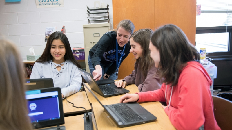 A group of students sitting at their desks with laptops. A teacher is standing pointing to a laptop.