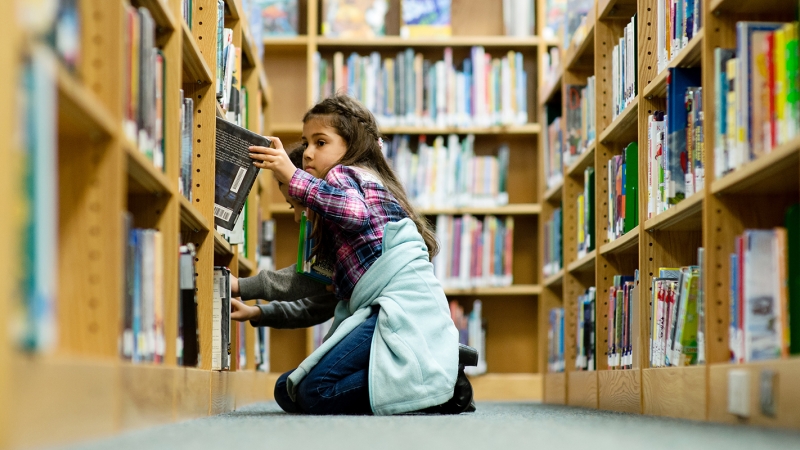 Photo of a female student looking for a book on a library shelf.