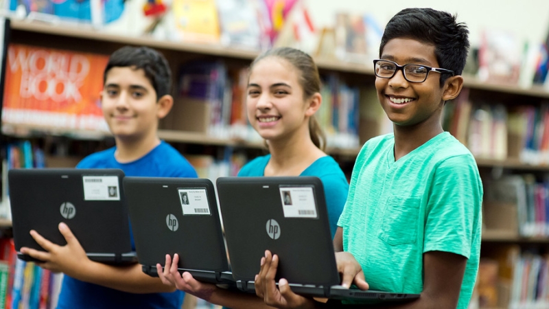 three students holding laptops in a school library