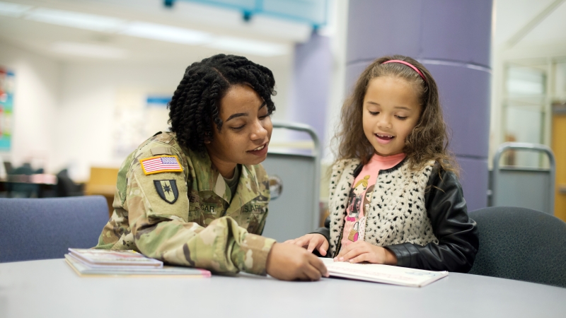 Photo of a mentor in a military uniform reading with a female elementary student