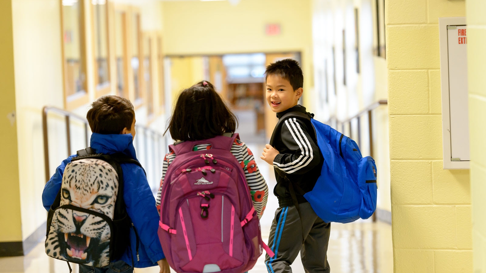 Elementary age students walking in a school hallway wearing backpacks. 